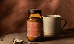 A jar labeled True Nightcap stands on a wooden surface next to a ceramic mug and a brass spoon filled with powder, against a warm brown, softly lit background.