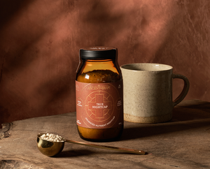 A jar labeled True Nightcap stands on a wooden surface next to a ceramic mug and a brass spoon filled with powder, against a warm brown, softly lit background.
