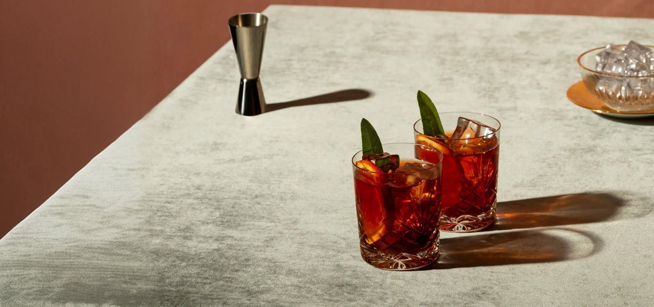 Two glasses of amber-colored cocktails with ice and green garnish sit on a light textured surface, next to a metal jigger and a plate of ice cubes in the background.