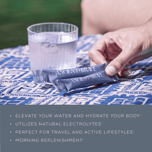 A hand holds an Ancient + Brave True Hydrate Sachet on a blue patterned cloth next to a glass of water. Text highlights its electrolyte-rich mineral blend for morning hydration, travel convenience, and support for active lifestyles.