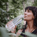 A woman with dark curly hair enjoys the outdoors, sipping from a clear bottle. Text: Start your day with Ancient + Brave True Hydrate Sachets—just add 6g to water for a refreshing mineral blend and natural coconut water flavor.