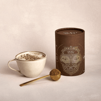 A cup of frothy hot chocolate sits beside a brown Ancient+Brave Cacao + Reishi container. A gold spoon with cocoa powder rests in front of the container, all set against a neutral background.
