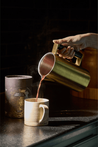 A hand pours a hot beverage from a gold-colored metal pitcher into a beige ceramic mug on a kitchen counter, with a decorative container labeled “CACAO + COLLAGEN” nearby. Steam rises gently from the mug.