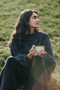 A woman with long dark hair sits on grass, wearing a navy blue sweater and holding a ceramic mug. She looks thoughtfully to the side, with sunlight shining on her and greenery in the background.