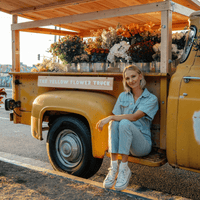 A woman in light denim sits on the step of a yellow flower truck filled with colorful bouquets in metal buckets, smiling at the camera on a sunny street. The trucks sign reads The Yellow Flower Truck.