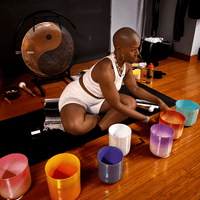 A woman in white workout attire sits on a mat indoors, surrounded by colorful crystal singing bowls. A large gong with a yin-yang symbol is behind her, and she appears to be preparing for a sound healing session.