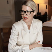 A woman with blonde hair in a low bun, wearing black glasses and a cream-colored blazer, sits at a table with a book, smiling slightly. A lamp and bed are visible in the softly lit background.