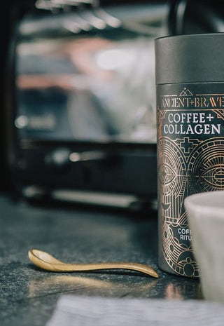 A coffee canister labeled Coffee + Collagen sits on a kitchen counter with a gray ceramic cup, a gold spoon, and a blurred espresso machine in the background.