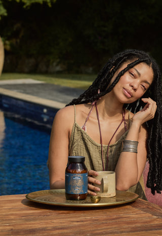 A woman with long braided hair sits by a pool, smiling softly while holding her face with one hand. She wears a sleeveless dress and jewelry, with a tray holding a supplement bottle, cup, and small container on the table in front of her.
