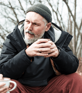 A man with a gray beard and wearing a green knit hat, dark jacket, and burgundy pants sits outdoors holding a mug with both hands, looking thoughtfully to his side. Trees without leaves are visible in the background.