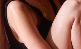 A close-up of a person sitting with knees drawn up, showing glistening, sweaty skin. The person wears a sleeveless black top, a gold necklace, and has a small tattoo on their upper arm.
