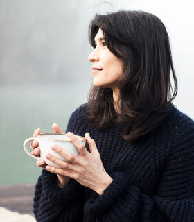 A woman with dark hair wearing a navy sweater holds a ceramic mug in both hands, looking thoughtfully into the distance. The background is softly blurred, creating a calm, serene atmosphere.