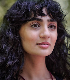 A woman with wavy dark hair and bangs is looking slightly upward. She has light makeup and a calm expression, with a blurred natural background behind her.