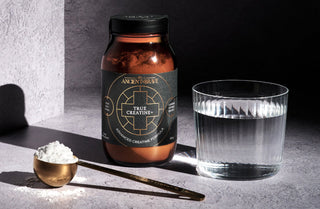 A jar labeled True Creatine stands next to a glass of water, a gold measuring spoon, and a mound of white powder on a textured gray surface with dramatic lighting and shadows.