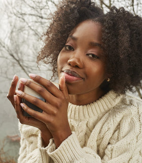 A woman with curly hair and a cream-colored sweater holds a mug with both hands, looking at the camera and smiling softly. Leafless tree branches are visible in the blurred background.