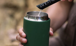 A person pours powder from an Ancient + Brave packet into a metal cup with a green insulated sleeve, held in their other hand. The background is blurred outdoors.