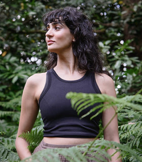 A woman with wavy dark hair wearing a black tank top stands among green ferns, looking to the side. Lush foliage and trees fill the background, creating a natural outdoor setting.