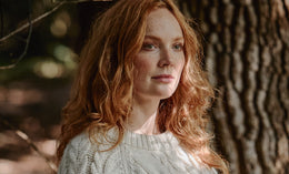 A woman with long red hair and fair skin, wearing a white knitted sweater, stands outdoors in front of a tree trunk with sunlight filtering through the branches.