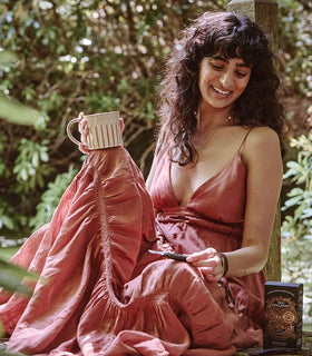 A woman in a flowing, rose-colored dress sits outdoors on a wooden deck, holding a mug and smiling while looking down. Next to her is a box labeled “Ceremonial Cacao.” Lush greenery surrounds her.