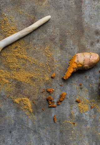 A wooden spoon with ground turmeric, fresh turmeric root, and cardamom pods are scattered on a rough metal surface.