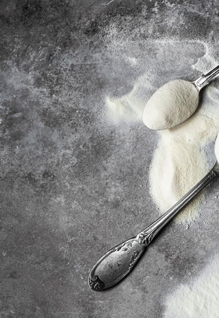 Three vintage silver spoons filled with white powder, possibly flour or powdered sugar, are scattered on a textured gray surface, with some powder spilled around them.