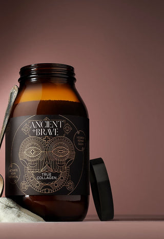 A brown glass jar labeled Ancient + Brave True Collagen stands on a surface with a spoon and a small mound of powder nearby, against a gradient brown background. The jars black lid rests beside it.