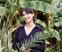 A woman with dark hair, wearing a navy blue top, stands smiling among green tropical plants and palm leaves in an outdoor garden.