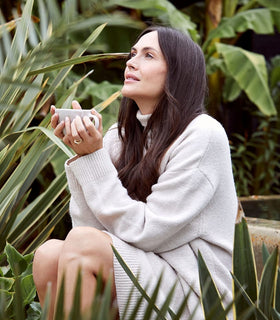 A woman with long dark hair sits among green plants, wearing a cozy white sweater dress. She holds a mug and looks up with a serene expression, surrounded by lush foliage.