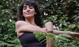 A woman with wavy dark hair wearing a sleeveless black top stretches her arm across her chest while standing outdoors, surrounded by green foliage and trees.