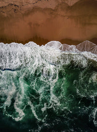 Aerial view of waves crashing onto a sandy beach, with white foam meeting deep green ocean water and a smooth, untouched shoreline above.