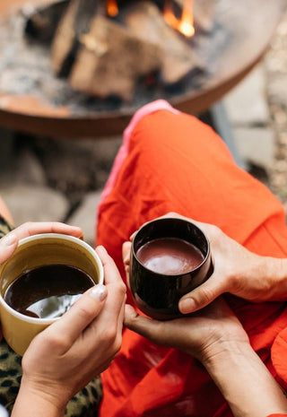 Two people sit near a fire pit, each holding a cup of hot drink. One wears a red outfit, the other wears animal print. Only their hands and part of their bodies are visible, creating a cozy, relaxed atmosphere.