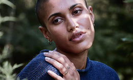 A person with closely shaved hair and a nose ring gazes softly at the camera, resting their hand on their shoulder. They wear a blue sweater and stand outdoors with greenery in the blurred background.