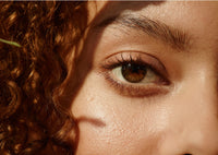 A close-up of a persons face, focusing on their right eye with long lashes and brown iris. Curly hair frames the side, and sunlight casts soft shadows across smooth, light-brown skin.