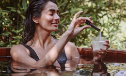 A woman with wet hair relaxes in an outdoor wooden tub, holding a glass of water and stirring it with a black stick labeled INVIVO. Green foliage surrounds her in the background.