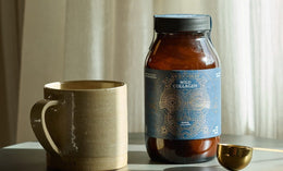 A brown ceramic mug, a dark brown jar labeled “Wild Collagen,” and a gold measuring spoon are arranged on a round table, with light curtains softly illuminated in the background.