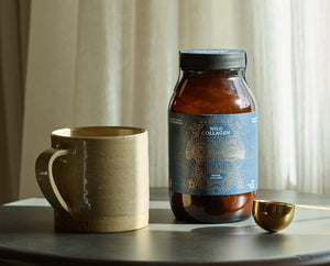 A brown ceramic mug, a dark brown jar labeled “Wild Collagen,” and a gold measuring spoon are arranged on a round table, with light curtains softly illuminated in the background.