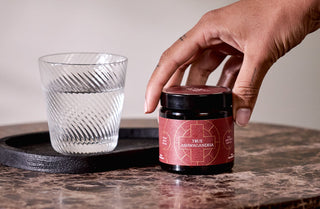 A hand reaches for a jar labeled True Ashwagandha on a marble table, next to a textured glass of water on a black tray.