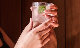 A person holds a glass of clear beverage with ice and a mint leaf, resting their arm on bent knees against a wooden background.